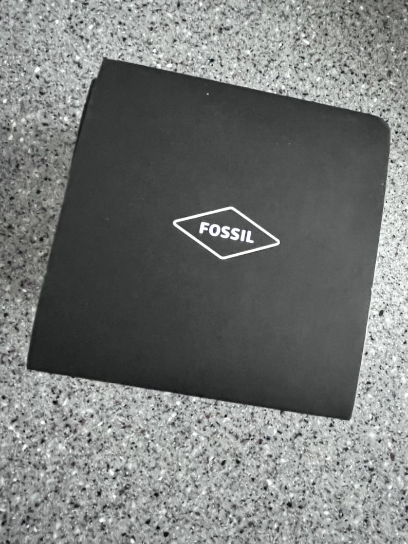 May include: A black square box with the brand name "FOSSIL" in a white diamond-shaped logo. The box is on a speckled gray surface.