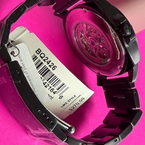 May include: A black Fossil watch with a skeleton dial, visible through the back. The watch has a black metal band and a tag with the style number BQ2426. The watch is displayed on a pink surface.