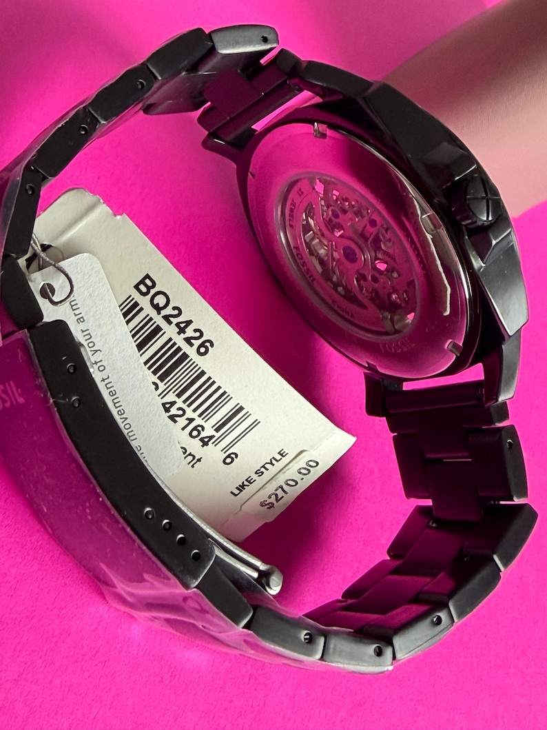 May include: A black Fossil watch with a skeleton dial, visible through the back. The watch has a black metal band and a tag with the style number BQ2426. The watch is displayed on a pink surface.