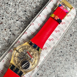 May include: A gold-toned Swatch watch with a transparent face, revealing the inner workings. It features a bright red leather strap and colorful accents on the band. The watch is presented in a clear plastic case with the text "Swatch Club".