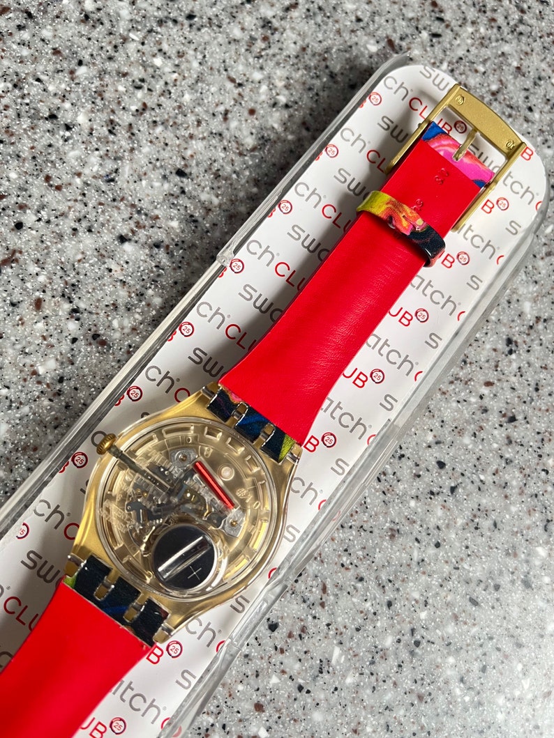 May include: A gold-toned Swatch watch with a transparent face, revealing the inner workings. It features a bright red leather strap and colorful accents on the band. The watch is presented in a clear plastic case with the text "Swatch Club".