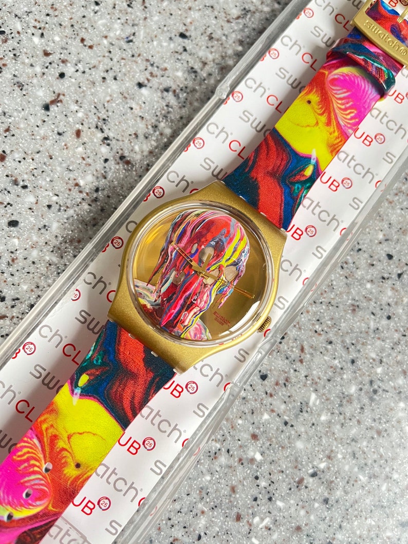 May include: Gold-toned wristwatch with a colorful abstract design on the face and strap. The watch face features a vibrant, multi-colored image. The strap displays a swirling pattern of red, yellow, and pink hues. The watch is in its original packaging.