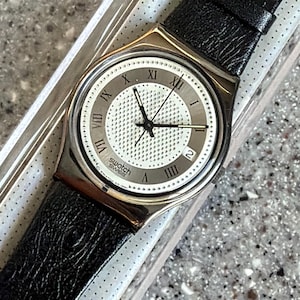 K&ouml;nnte beinhalten: Eine Swatch Swiss Uhr mit einem schwarzen Lederarmband. Das Zifferblatt der Uhr ist wei&szlig; mit schwarzen Zeigern und r&ouml;mischen Ziffern. Die Uhr befindet sich in der Originalverpackung.