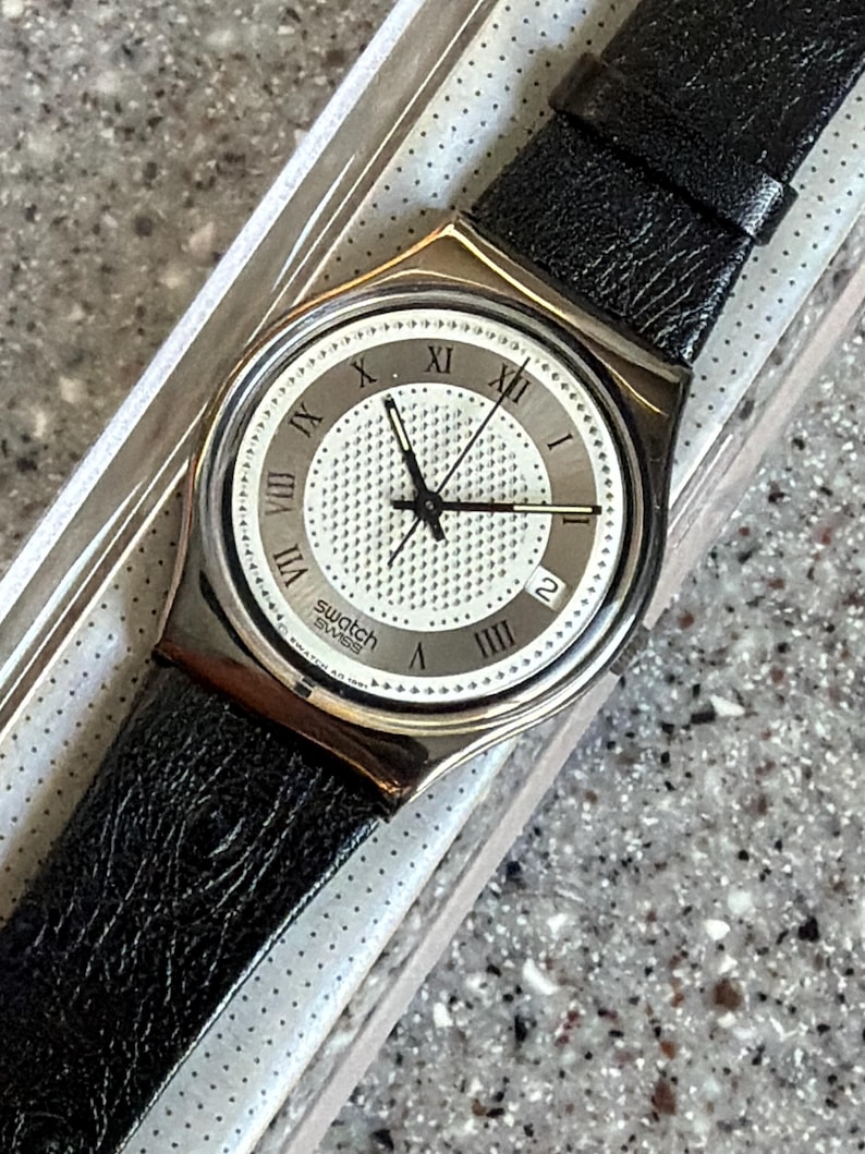 K&ouml;nnte beinhalten: Eine Swatch Swiss Uhr mit einem schwarzen Lederarmband. Das Zifferblatt der Uhr ist wei&szlig; mit schwarzen Zeigern und r&ouml;mischen Ziffern. Die Uhr befindet sich in der Originalverpackung.