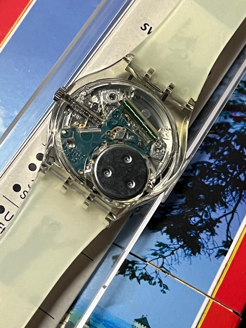 Op de afbeelding: Een transparant plastic horloge met een witte band. De wijzerplaat van het horloge is transparant, waardoor het innerlijk van het horloge zichtbaar is. Het horloge ligt op een wit oppervlak.