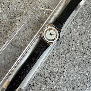 K&ouml;nnte beinhalten: Eine Vintage-Armbanduhr mit schwarzem Lederarmband und silberfarbenem Geh&auml;use. Das Zifferblatt ist wei&szlig; mit schwarzen Zeigern und r&ouml;mischen Ziffern. Die Uhr befindet sich in ihrer originalen transparenten Kunststoffverpackung.
