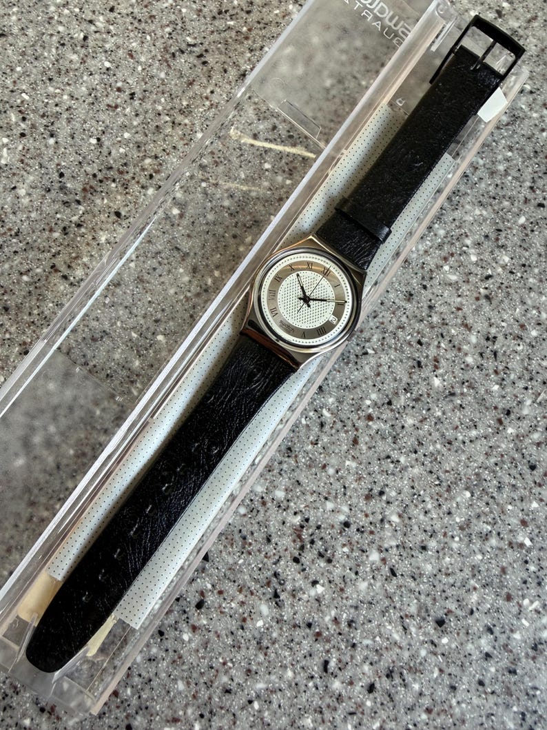 K&ouml;nnte beinhalten: Eine Vintage-Armbanduhr mit schwarzem Lederarmband und silberfarbenem Geh&auml;use. Das Zifferblatt ist wei&szlig; mit schwarzen Zeigern und r&ouml;mischen Ziffern. Die Uhr befindet sich in ihrer originalen transparenten Kunststoffverpackung.