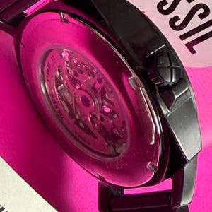 May include: Close-up of a black Fossil watch with a visible skeleton movement. The watch has a black metal band and a pink background. The watch face is visible, showcasing the intricate inner workings.