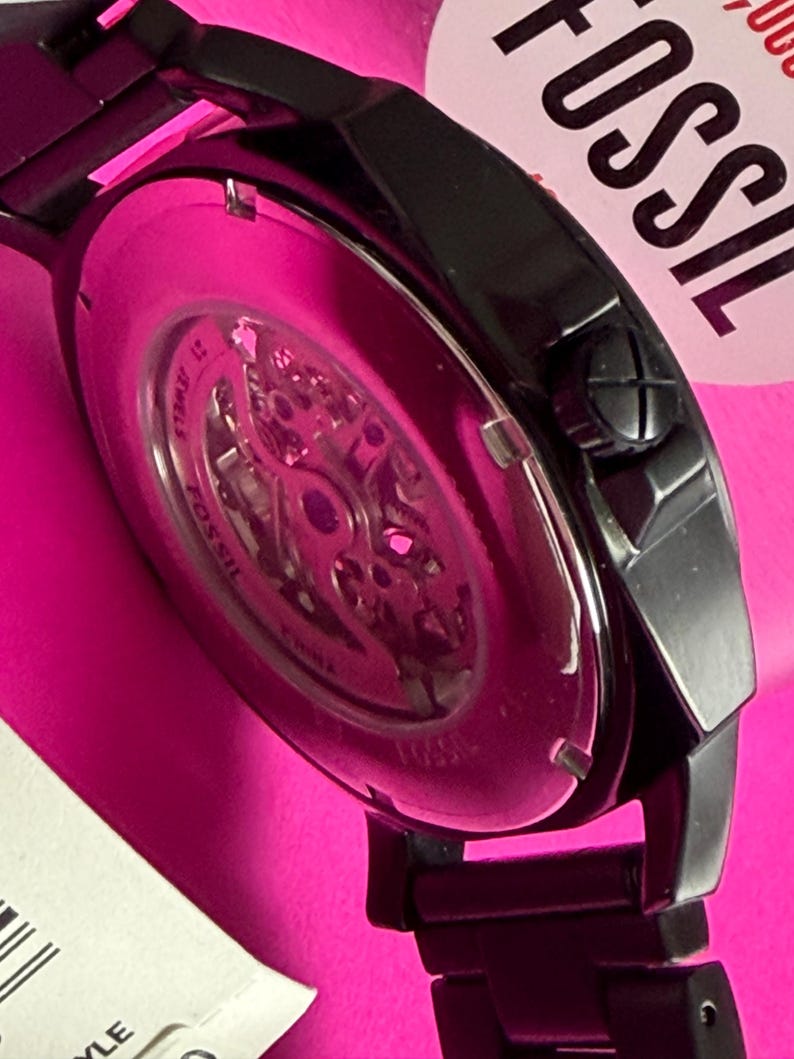 May include: Close-up of a black Fossil watch with a visible skeleton movement. The watch has a black metal band and a pink background. The watch face is visible, showcasing the intricate inner workings.