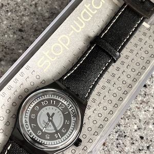 Puede incluir: Un reloj Swatch negro con una esfera blanca y agujas negras. El reloj tiene una correa de cuero negro con costuras blancas. El reloj est&aacute; en una caja de pl&aacute;stico transparente con las palabras "Stop-watch" impresas en ella.