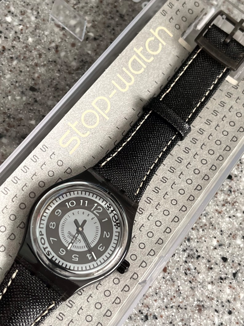 Puede incluir: Un reloj Swatch negro con una esfera blanca y agujas negras. El reloj tiene una correa de cuero negro con costuras blancas. El reloj est&aacute; en una caja de pl&aacute;stico transparente con las palabras "Stop-watch" impresas en ella.