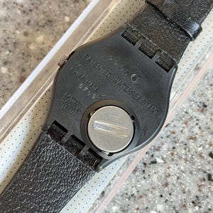 K&ouml;nnte beinhalten: Nahaufnahme der R&uuml;ckseite einer schwarzen Armbanduhr mit strukturiertem Lederarmband. Das Zifferblatt der Uhr ist schwarz mit silbernen Akzenten. Auf der Uhr steht "PATENTED WATERRESISTANT" und "SWISS MADE".