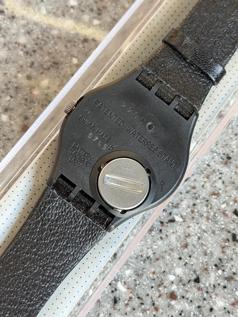 K&ouml;nnte beinhalten: Nahaufnahme der R&uuml;ckseite einer schwarzen Armbanduhr mit strukturiertem Lederarmband. Das Zifferblatt der Uhr ist schwarz mit silbernen Akzenten. Auf der Uhr steht "PATENTED WATERRESISTANT" und "SWISS MADE".