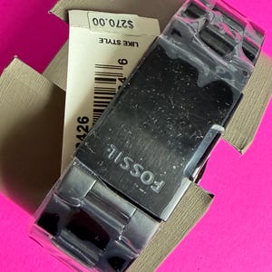 May include: A Fossil watch with a black face and silver band is displayed in its original box. The watch is partially covered in plastic. The watch's band is made of metal. The watch is in a tan box on a pink background.