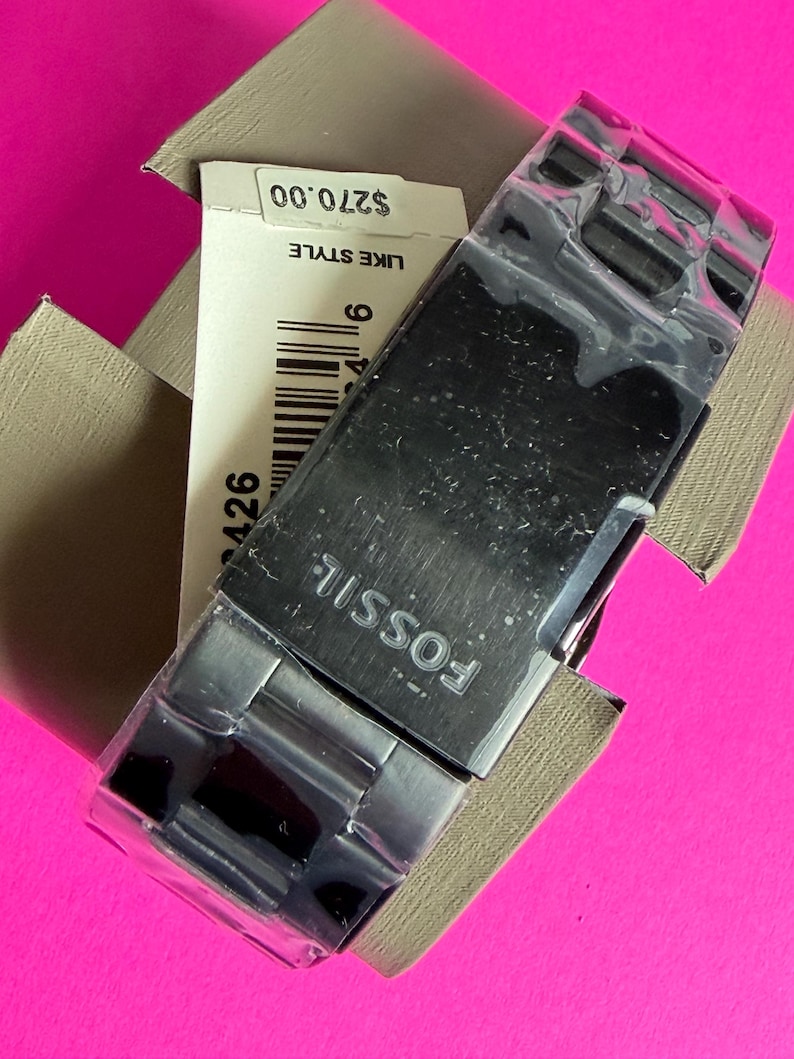 May include: A Fossil watch with a black face and silver band is displayed in its original box. The watch is partially covered in plastic. The watch's band is made of metal. The watch is in a tan box on a pink background.