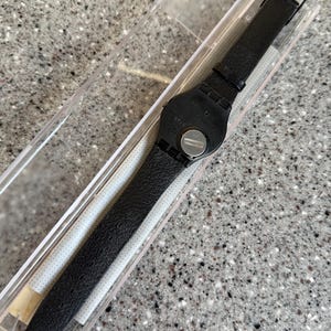 K&ouml;nnte beinhalten: Eine schwarze Swatch-Uhr mit strukturiertem Lederarmband wird in einer transparenten Kunststoffbox pr&auml;sentiert. Die Uhr hat ein schwarzes Zifferblatt und eine silberne Batterieabdeckung. Das Armband ist etwa 23 cm lang.