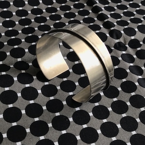 Op de afbeelding: Een zilveren metalen armband met een gladde, gepolijste afwerking. De armband is open en heeft een licht gebogen vorm.