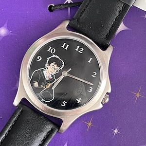 May include: A black leather wristwatch with a silver face featuring a Harry Potter character illustration. The watch face has black numbers and hands. The watch is on a purple background with stars.