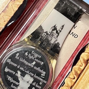Op de afbeelding: Een wit plastic horloge met een zwart-witte afbeelding van Neuschwanstein Castle op de wijzerplaat. De wijzerplaat van het horloge heeft ook de tekst "Expose to light source" in het Engels, Frans, Duits, Italiaans en Spaans.