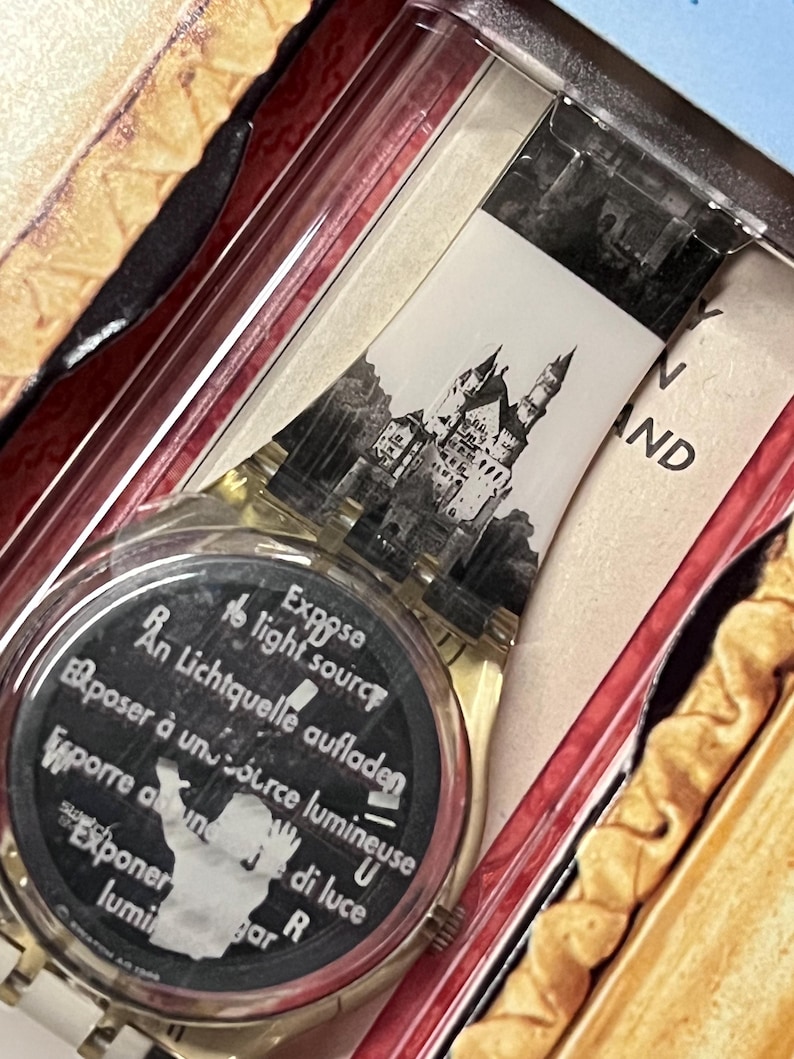 Op de afbeelding: Een wit plastic horloge met een zwart-witte afbeelding van Neuschwanstein Castle op de wijzerplaat. De wijzerplaat van het horloge heeft ook de tekst "Expose to light source" in het Engels, Frans, Duits, Italiaans en Spaans.