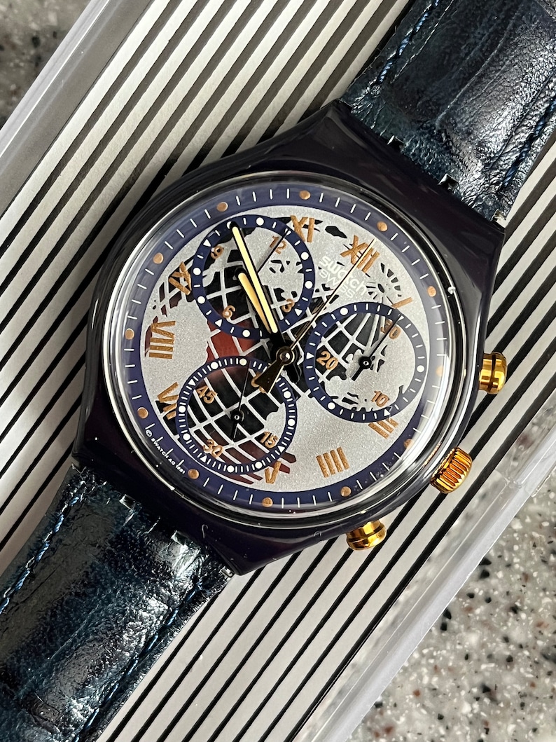 May include: A blue and silver Swatch watch with a chronograph and a world map design on the face. The watch has a blue leather strap.