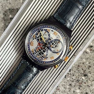 May include: A blue Swatch watch with a silver face and gold accents. The watch has a chronograph function and a world time display. The watch is in its original box.