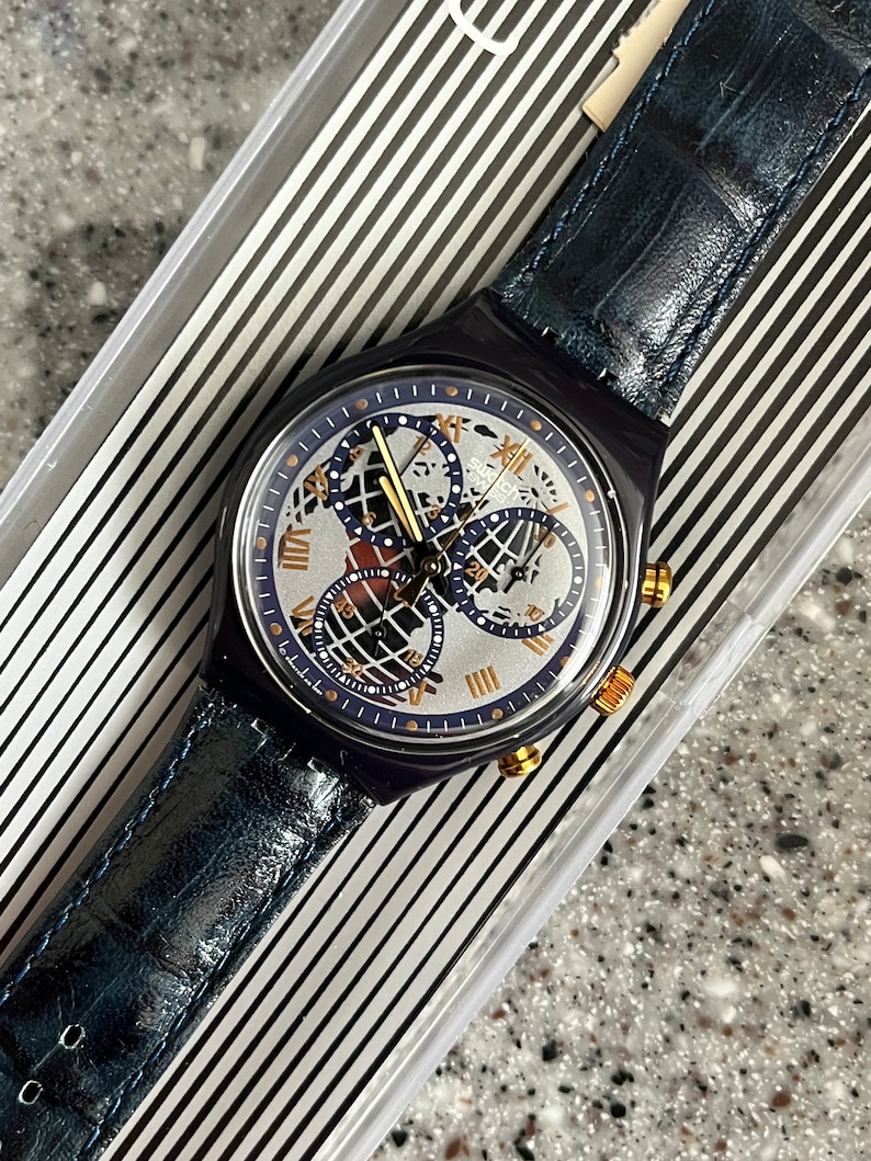 May include: A blue Swatch watch with a silver face and gold accents. The watch has a chronograph function and a world time display. The watch is in its original box.