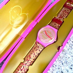 May include: A pink Swatch watch with a gold dial and a patterned strap, presented in a pink and gold case. The watch face displays the words "WORLD COMMUNICATION FUTURE". The strap features a floral design.