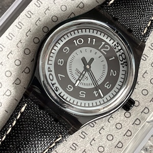 Puede incluir: Un reloj Swatch negro con una correa de cuero negro y una esfera blanca. La esfera del reloj tiene un anillo exterior negro con n&uacute;meros blancos y un c&iacute;rculo interior blanco m&aacute;s peque&ntilde;o con agujas negras y la palabra "segundos" impresa en &eacute;l. La esfera del reloj tambi&eacute;n tiene la palabra "STOP" impresa en &eacute;l en un c&iacute;rculo negro.