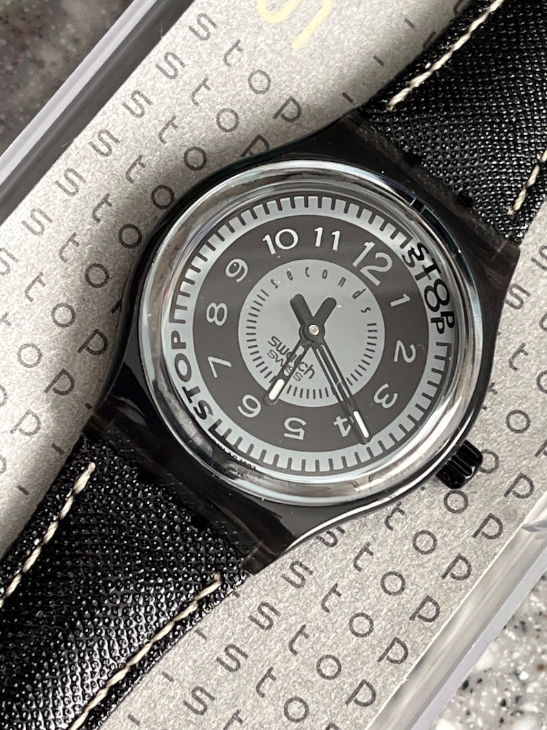 Puede incluir: Un reloj Swatch negro con una correa de cuero negro y una esfera blanca. La esfera del reloj tiene un anillo exterior negro con n&uacute;meros blancos y un c&iacute;rculo interior blanco m&aacute;s peque&ntilde;o con agujas negras y la palabra "segundos" impresa en &eacute;l. La esfera del reloj tambi&eacute;n tiene la palabra "STOP" impresa en &eacute;l en un c&iacute;rculo negro.