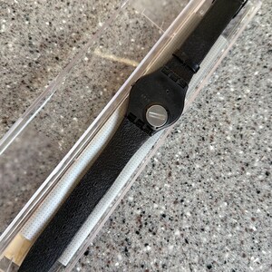 K&ouml;nnte beinhalten: Eine schwarze Armbanduhr mit Lederarmband wird in einer transparenten Kunststoffbox pr&auml;sentiert. Die Uhr hat ein schwarzes Zifferblatt und eine silberne Schnalle. Das Armband ist strukturiert und scheint aus Leder zu sein. Die Uhr ist neu und ungetragen.