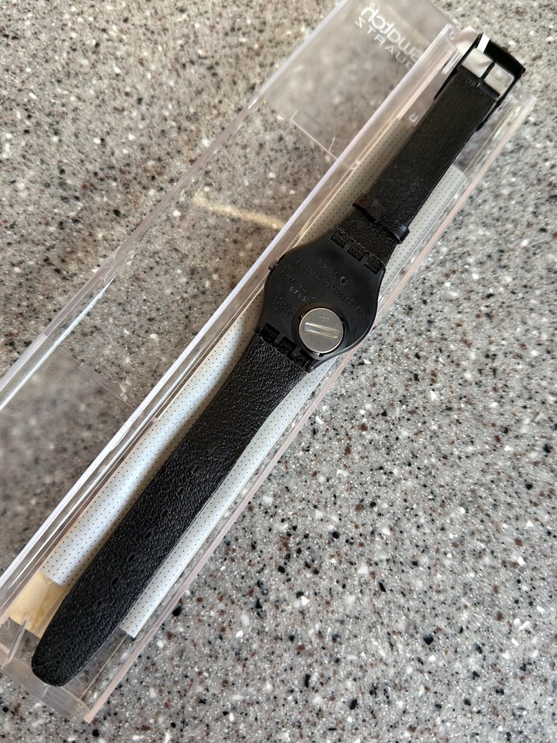 K&ouml;nnte beinhalten: Eine schwarze Armbanduhr mit Lederarmband wird in einer transparenten Kunststoffbox pr&auml;sentiert. Die Uhr hat ein schwarzes Zifferblatt und eine silberne Schnalle. Das Armband ist strukturiert und scheint aus Leder zu sein. Die Uhr ist neu und ungetragen.