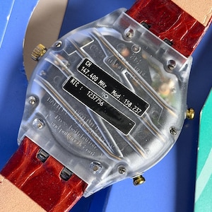 May include: A clear, transparent watch with a red leather band. The watch back displays technical information, including "CH 147.400 MHz" and "RIC: 1237756". The band is a deep red with a textured pattern.