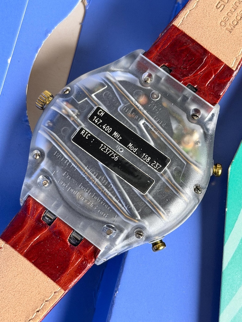 May include: A clear, transparent watch with a red leather band. The watch back displays technical information, including "CH 147.400 MHz" and "RIC: 1237756". The band is a deep red with a textured pattern.