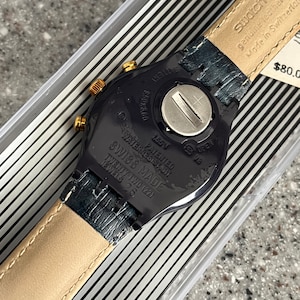 May include: A black Swatch watch with a gold-tone bezel and a black leather strap. The watch face is black with white markings and the text "WATER RESISTANT", "PATENTED", "SWISS MADE", "TWENTY TWO (22)", and "JEWELS 26" is printed on the back. The watch is in a clear plastic box with a black and white striped background.