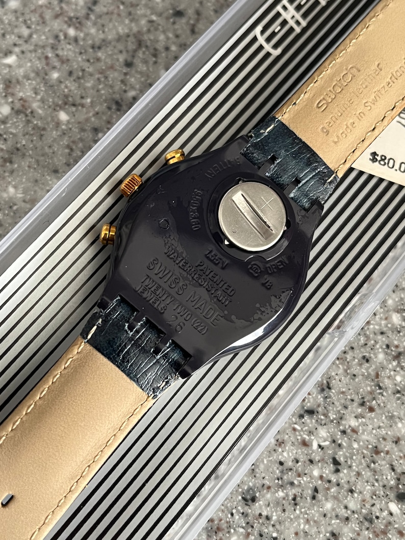 May include: A black Swatch watch with a gold-tone bezel and a black leather strap. The watch face is black with white markings and the text "WATER RESISTANT", "PATENTED", "SWISS MADE", "TWENTY TWO (22)", and "JEWELS 26" is printed on the back. The watch is in a clear plastic box with a black and white striped background.