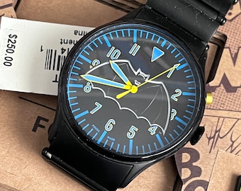 Puede incluir: Un reloj de pulsera negro con una esfera azul y un diseño de murciélago. El reloj tiene detalles amarillos y las palabras "Fossil Made" en la esfera.