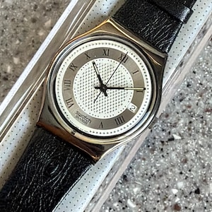 K&ouml;nnte beinhalten: Eine Swatch-Uhr mit silberfarbenem Geh&auml;use und schwarzem Lederarmband. Das Zifferblatt der Uhr ist wei&szlig; mit schwarzen r&ouml;mischen Ziffern und Zeigern. Die Uhr hat auch eine Datumsanzeige bei 3 Uhr.
