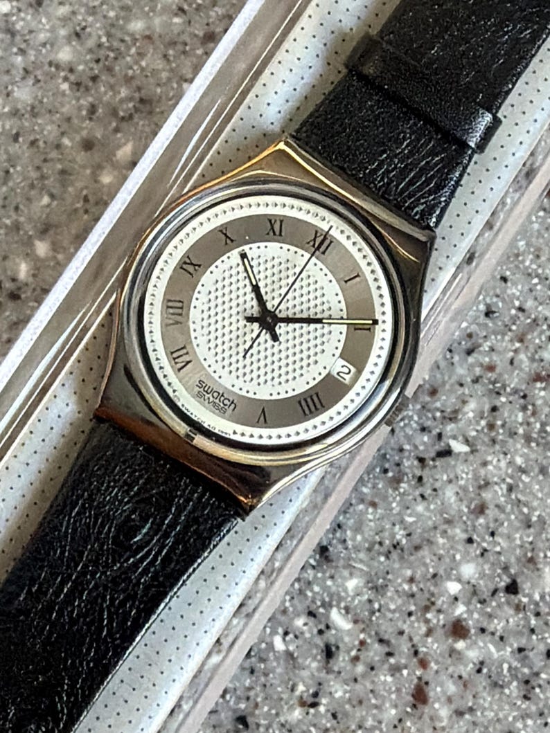 K&ouml;nnte beinhalten: Eine Swatch-Uhr mit silberfarbenem Geh&auml;use und schwarzem Lederarmband. Das Zifferblatt der Uhr ist wei&szlig; mit schwarzen r&ouml;mischen Ziffern und Zeigern. Die Uhr hat auch eine Datumsanzeige bei 3 Uhr.