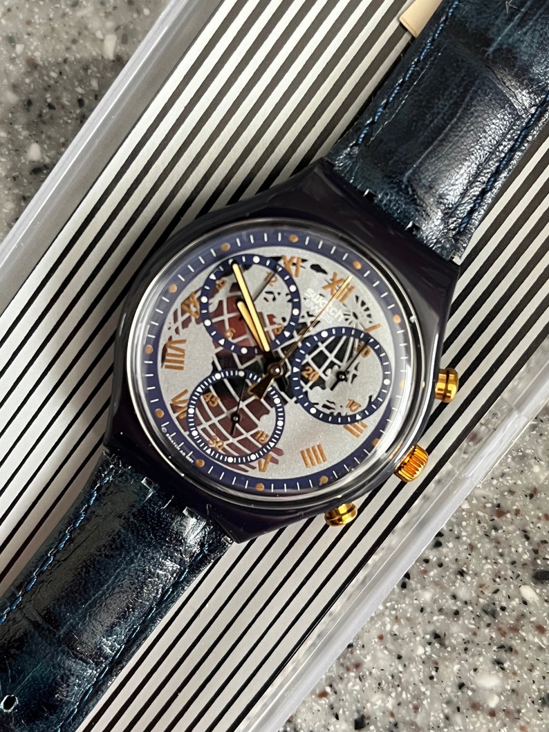 May include: A blue and silver Swatch watch with a chronograph and a world map design on the face. The watch has a blue leather strap and gold accents.