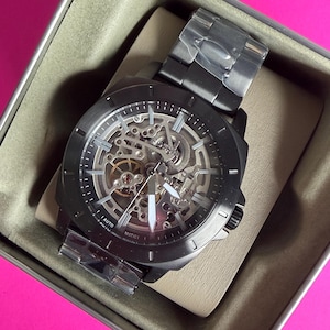 May include: A black skeleton watch with a stainless steel band, displayed in a gray watch box. The watch face reveals the inner workings of the automatic movement. The watch is new and still has protective plastic on the band.