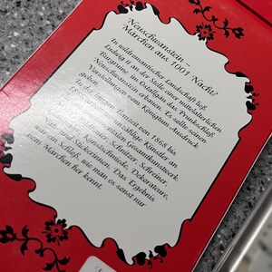 Op de afbeelding: Een rode doos met een wit label waarop staat "Neuschwanstein - M&auml;rchen aus 1001 Nacht?" Het label beschrijft het kasteel Neuschwanstein in Duitsland, gebouwd door koning Ludwig II. De doos heeft een prijskaartje met de tekst "DM 115.00".