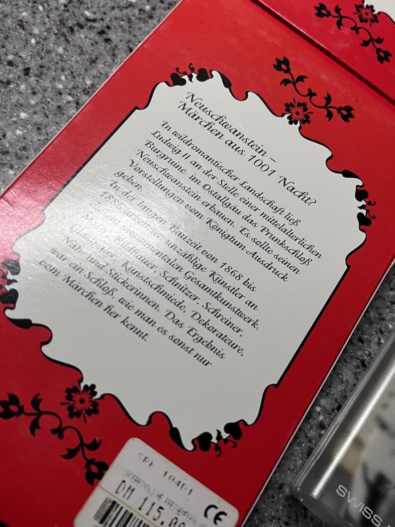Op de afbeelding: Een rode doos met een wit label waarop staat "Neuschwanstein - M&auml;rchen aus 1001 Nacht?" Het label beschrijft het kasteel Neuschwanstein in Duitsland, gebouwd door koning Ludwig II. De doos heeft een prijskaartje met de tekst "DM 115.00".