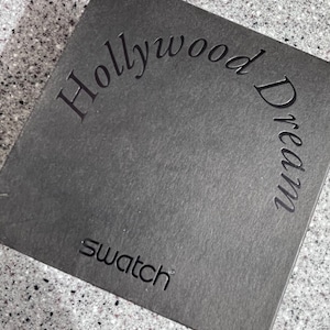 May include: A black box with the text "Hollywood Dream" in a curved font and the word "Swatch" in a smaller font below.