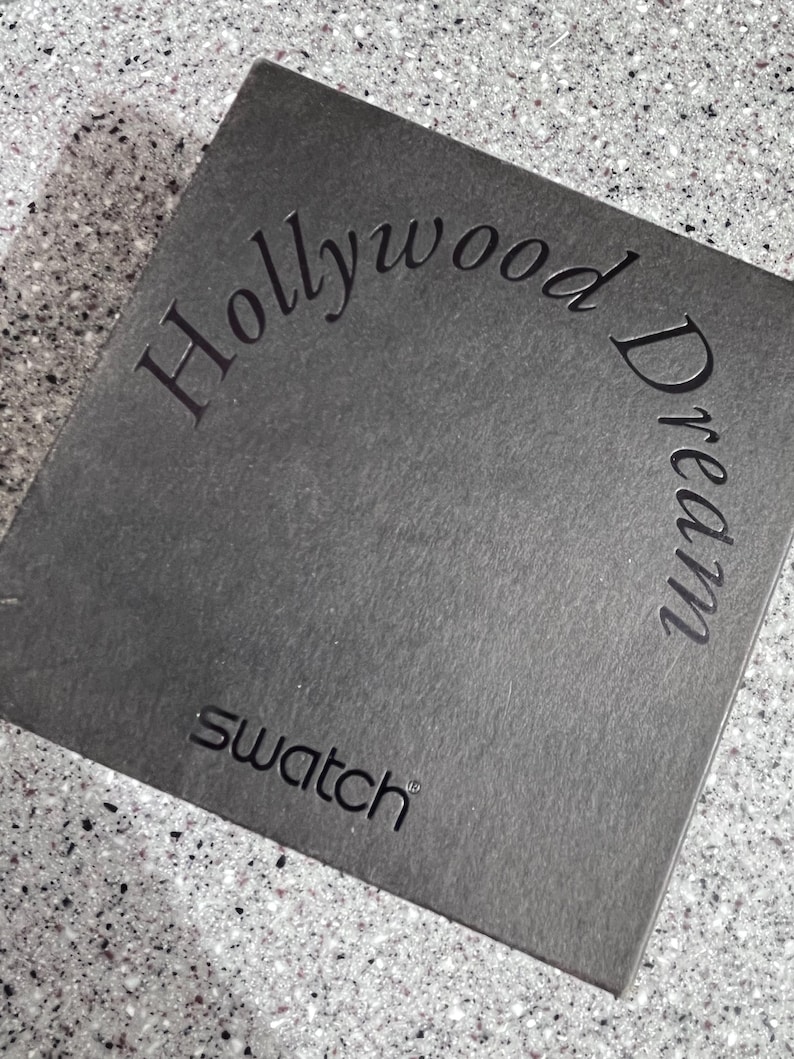 May include: A black box with the text "Hollywood Dream" in a curved font and the word "Swatch" in a smaller font below.
