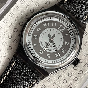 Puede incluir: Un reloj de pulsera negro con una correa de cuero negro y costuras blancas. La esfera del reloj es negra con n&uacute;meros blancos y una aguja de segundos plateada. La esfera del reloj tambi&eacute;n tiene el texto "Swatch Swiss" y "Stop" impreso en ella.