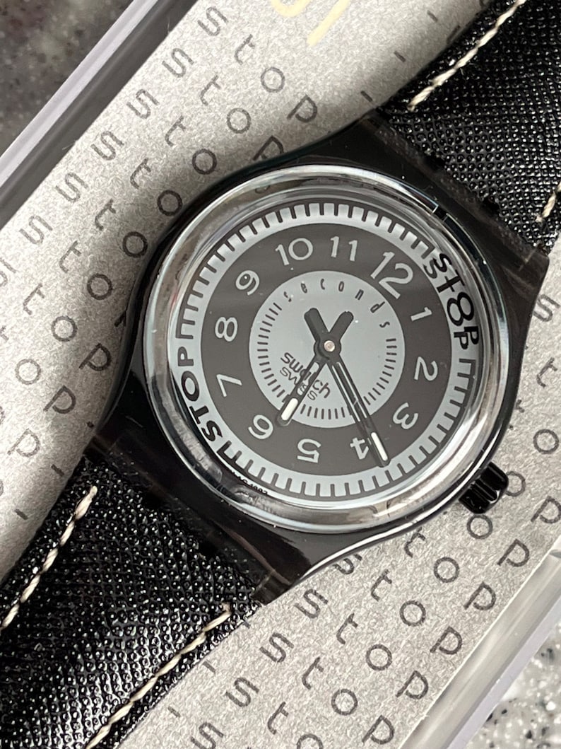 Puede incluir: Un reloj de pulsera negro con una correa de cuero negro y costuras blancas. La esfera del reloj es negra con n&uacute;meros blancos y una aguja de segundos plateada. La esfera del reloj tambi&eacute;n tiene el texto "Swatch Swiss" y "Stop" impreso en ella.