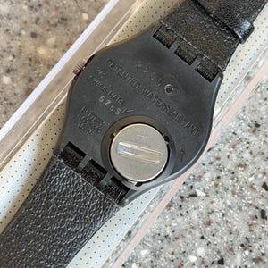 K&ouml;nnte beinhalten: Schwarze Armbanduhr mit strukturiertem Lederarmband. Die R&uuml;ckseite der Uhr ist schwarz mit dem Text "PATENTED WATERRESISTANT" und "SWISS MADE". Die Uhr hat ein silbernes Batteriefach und ein schwarzes Armband.