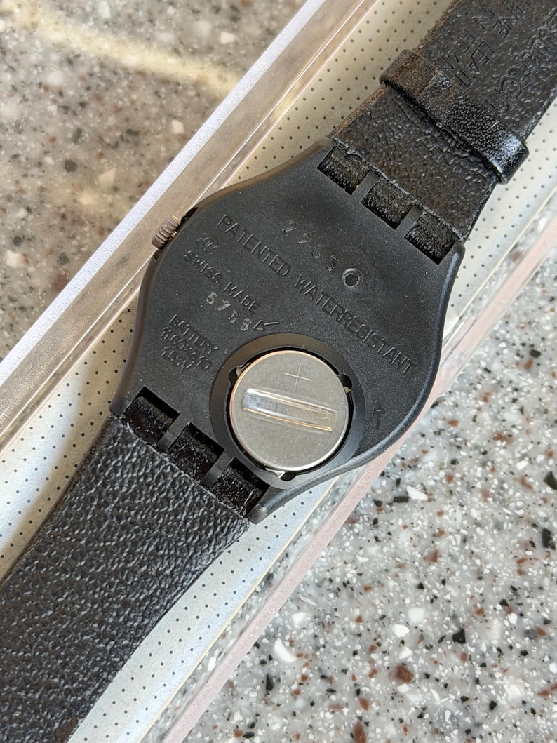 K&ouml;nnte beinhalten: Schwarze Armbanduhr mit strukturiertem Lederarmband. Die R&uuml;ckseite der Uhr ist schwarz mit dem Text "PATENTED WATERRESISTANT" und "SWISS MADE". Die Uhr hat ein silbernes Batteriefach und ein schwarzes Armband.