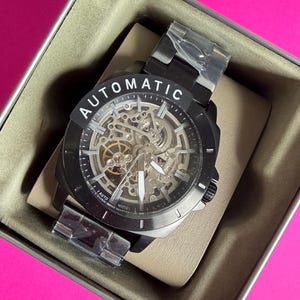 May include: Black automatic watch with a skeleton dial, visible gears, and white hands. The watch is in a metal case with the word "AUTOMATIC" on the bezel. The watch is new, with protective plastic on the band.